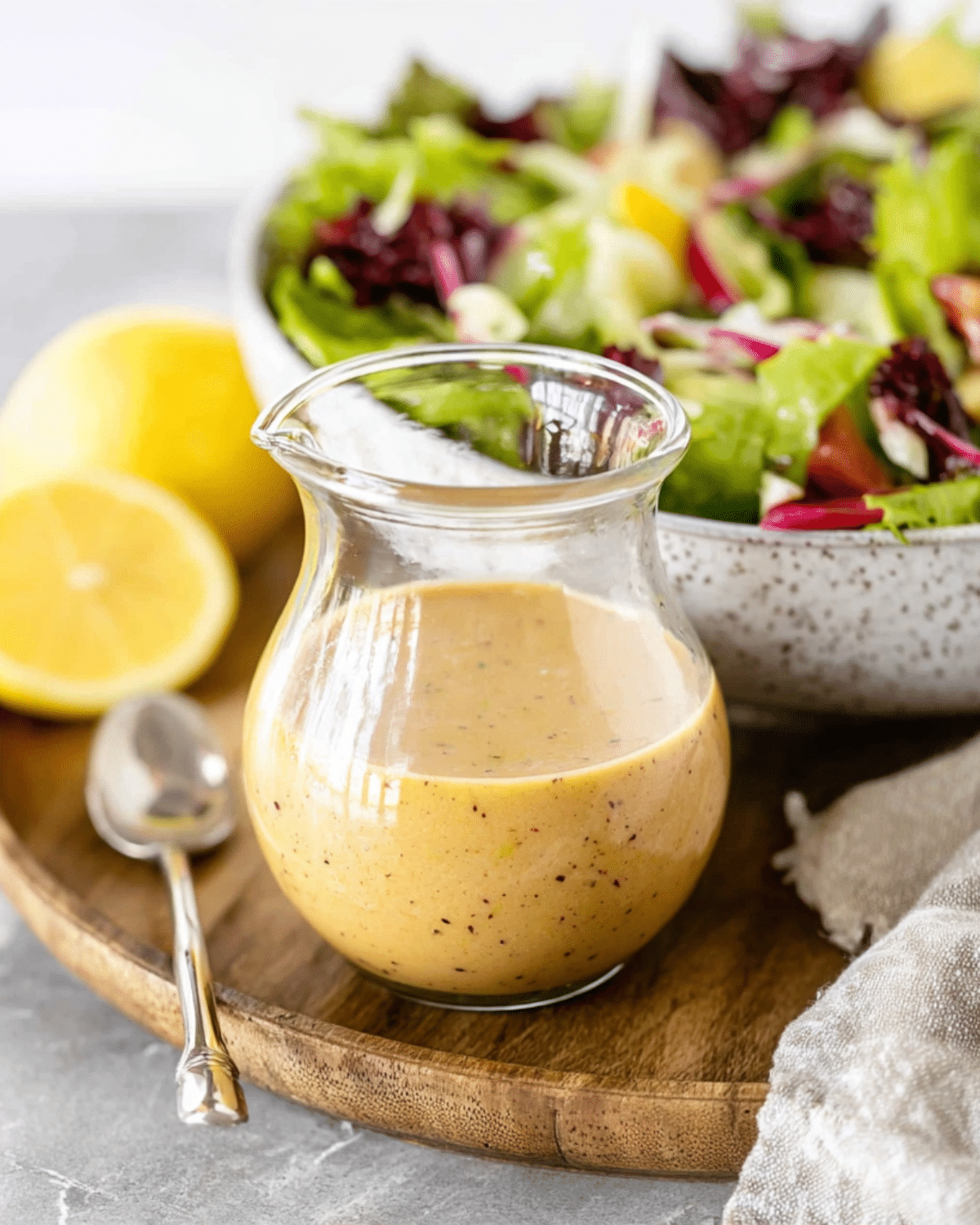 Glass jar of Lemon Vinaigrette Salad Dressing placed on a wooden tray with fresh lemon halves and a bowl of mixed salad in the background.