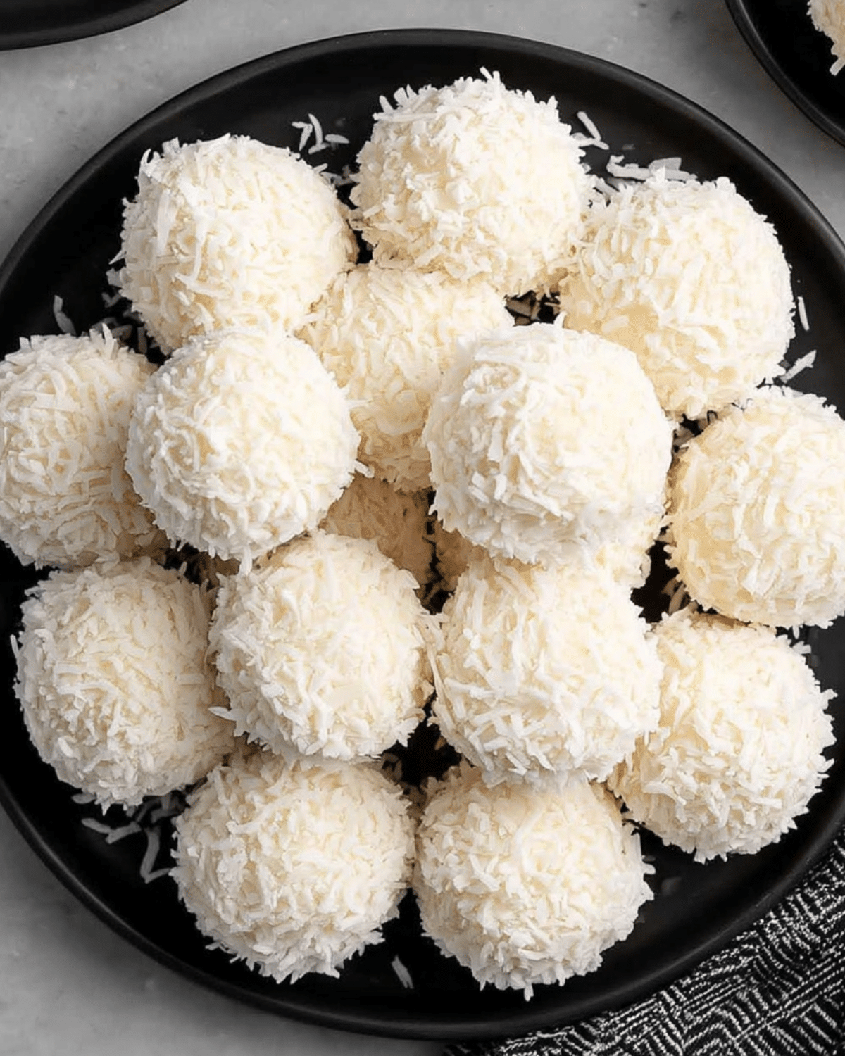 A plate of no bake coconut snowballs, coated in shredded coconut and arranged neatly on a black plate.