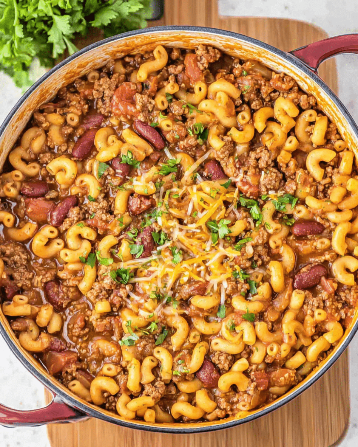 A pot of one pot mac chili filled with macaroni, ground beef, kidney beans, tomatoes, and topped with shredded cheese and fresh herbs.