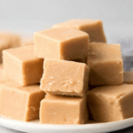 Smooth and creamy peanut butter fudge squares stacked on a white plate with a soft background.