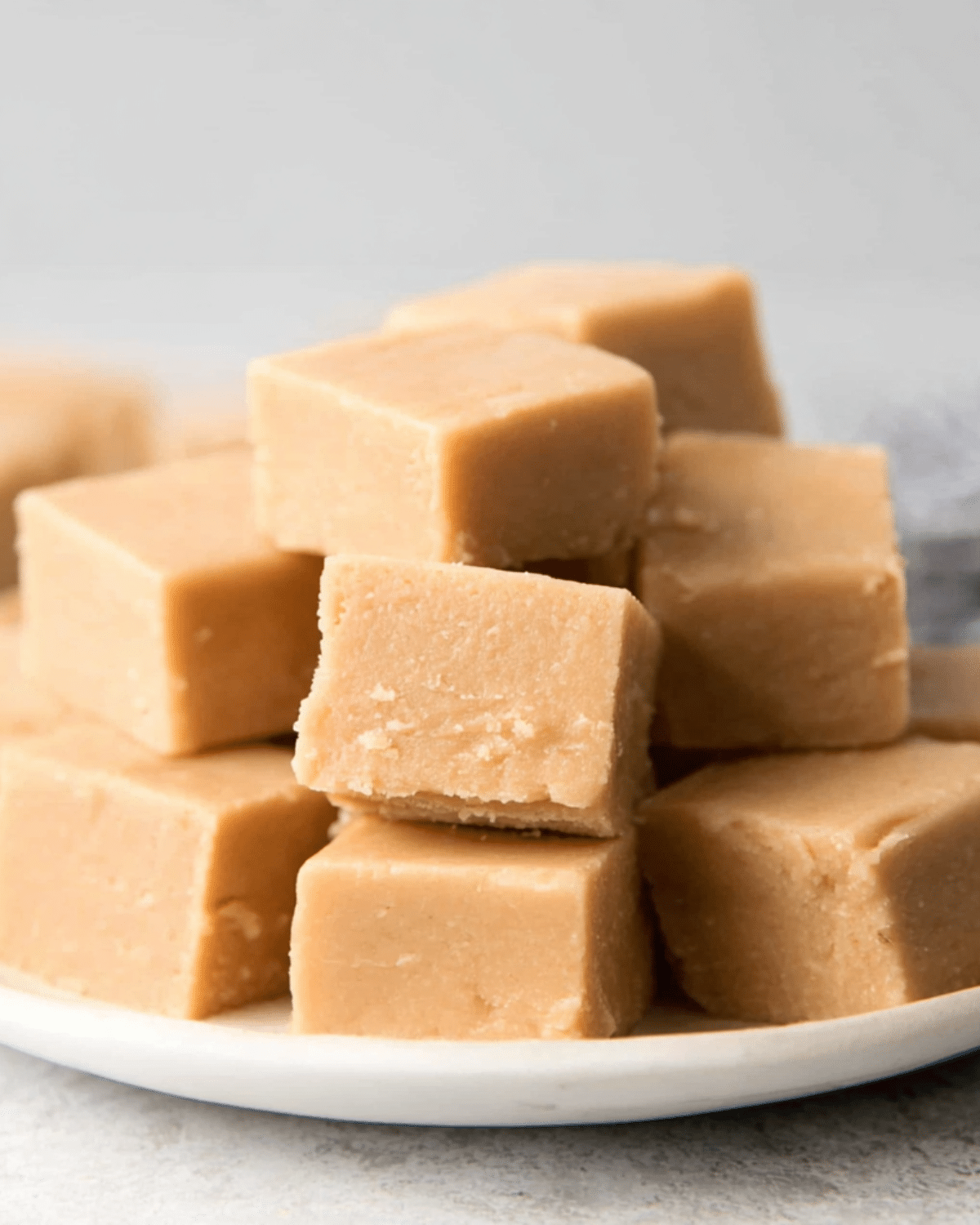 Smooth and creamy peanut butter fudge squares stacked on a white plate with a soft background.