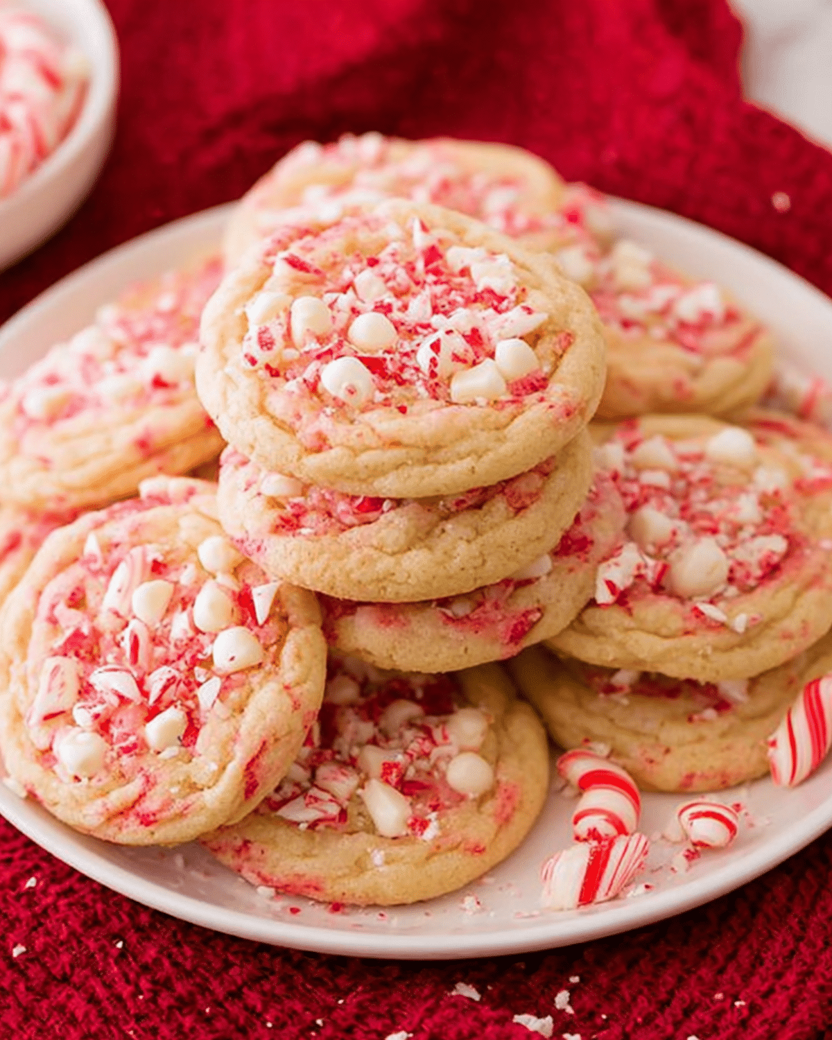 A plate of peppermint cookies topped with crushed candy canes and white chocolate chips, displayed on a red cloth.