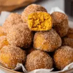 Plate of pumpkin donut holes coated in cinnamon sugar, with one cut open to show the soft, moist pumpkin interior.
