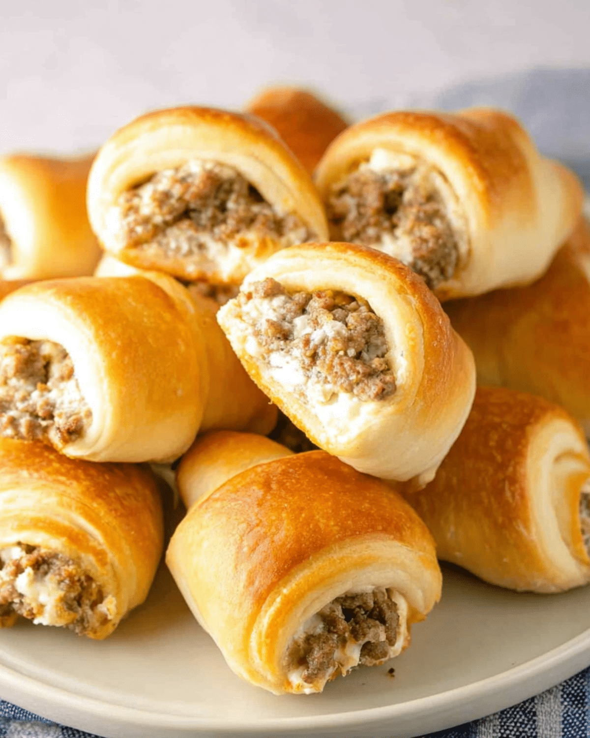 Golden sausage cream cheese crescent rolls stacked on a plate, with flaky pastry and a savory filling visible.