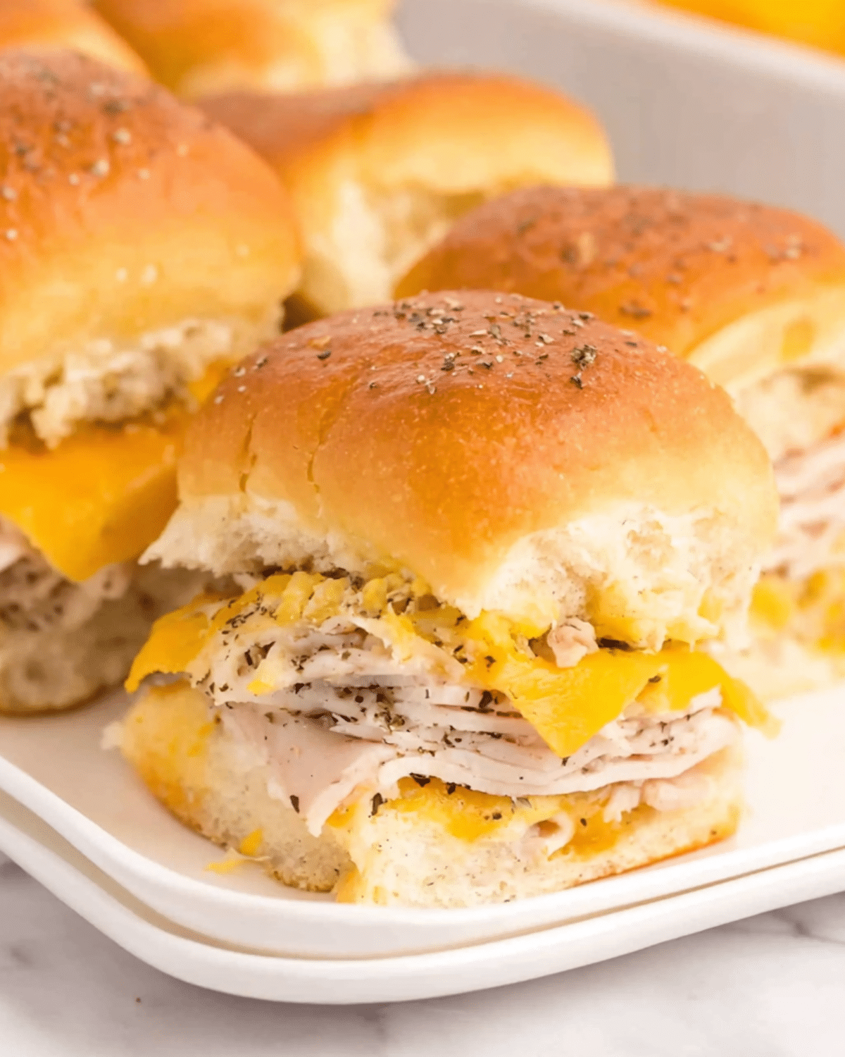 Close-up of turkey sliders with cheddar cheese on soft, golden buns, topped with herbs and served on a white plate.