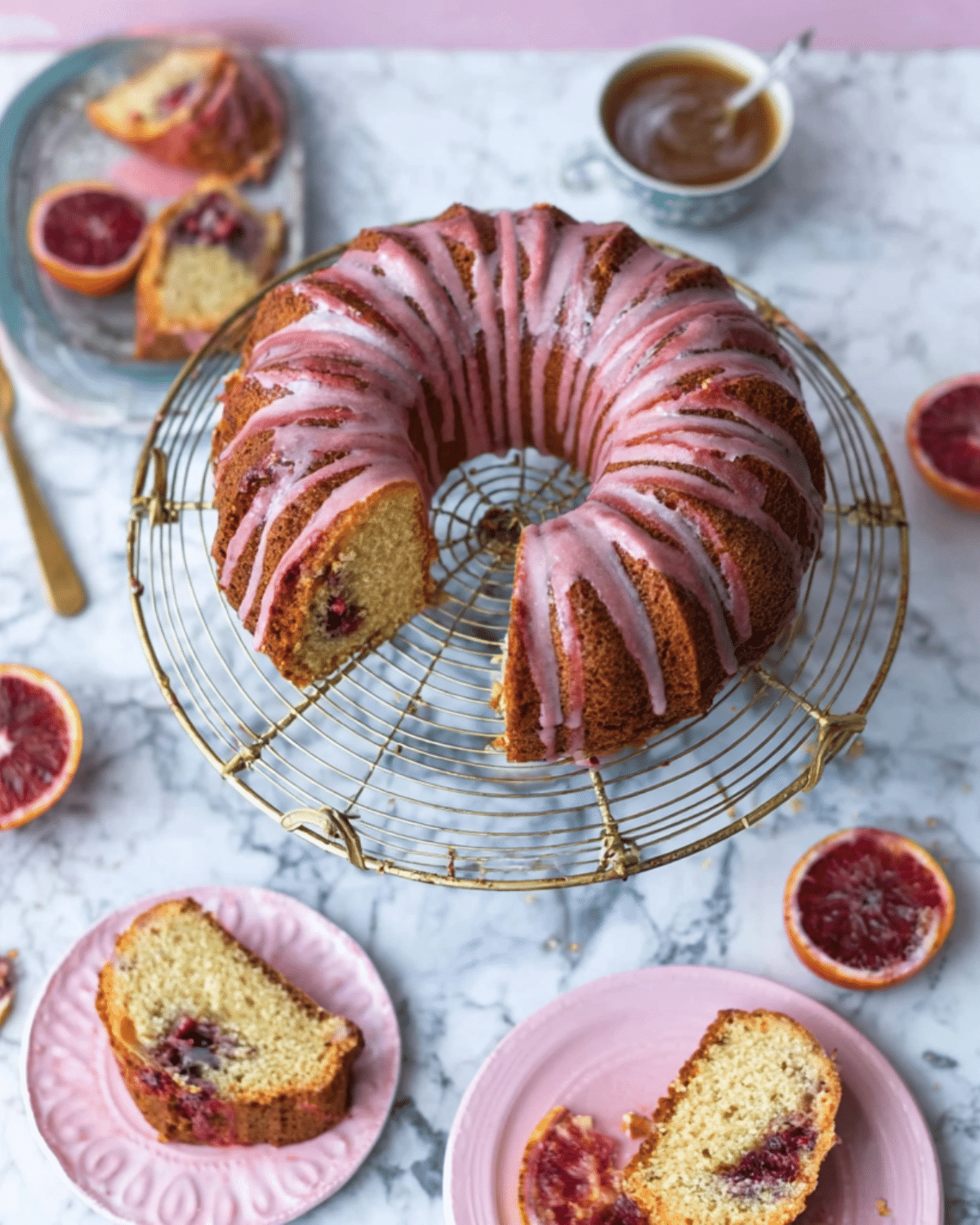 Blood orange coffee cake with pink glaze on a wire rack, surrounded by sliced cake pieces and fresh blood oranges.