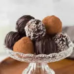 A glass dessert stand filled with assorted chocolate rum balls coated in cocoa powder, shredded coconut, and chocolate drizzle.