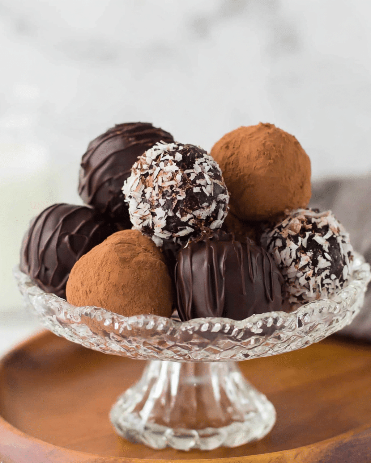 A glass dessert stand filled with assorted chocolate rum balls coated in cocoa powder, shredded coconut, and chocolate drizzle.
