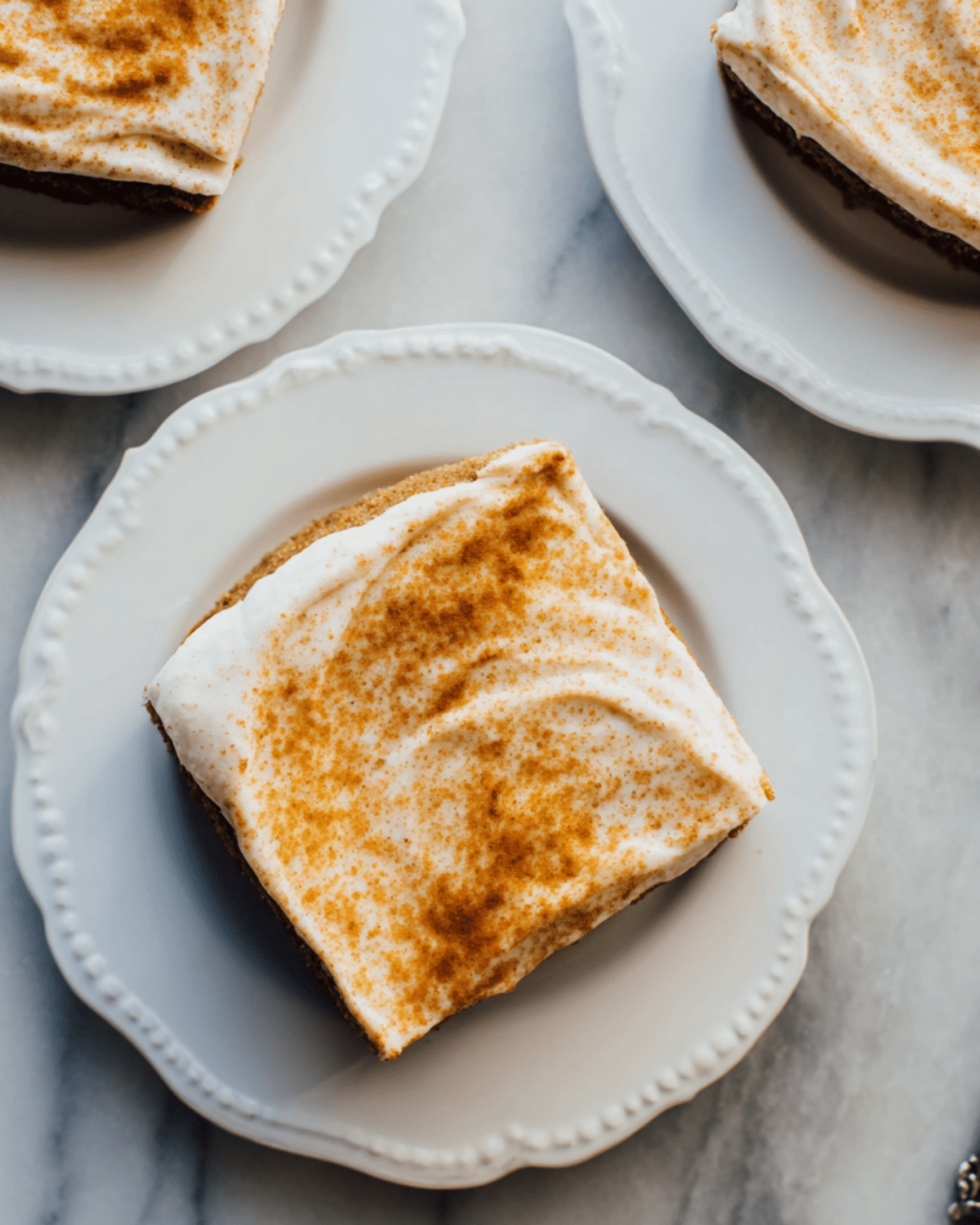 Slice of gingerbread cake with luscious cream cheese frosting, dusted with cinnamon, served on a white plate.