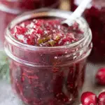 A glass jar filled with vibrant hot pepper cranberry jam, with visible pepper and cranberry pieces, and a spoon inside the jar.