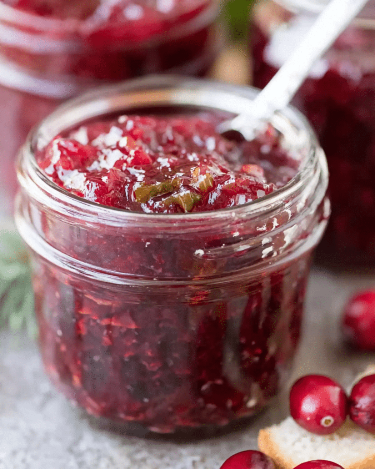 A glass jar filled with vibrant hot pepper cranberry jam, with visible pepper and cranberry pieces, and a spoon inside the jar.
