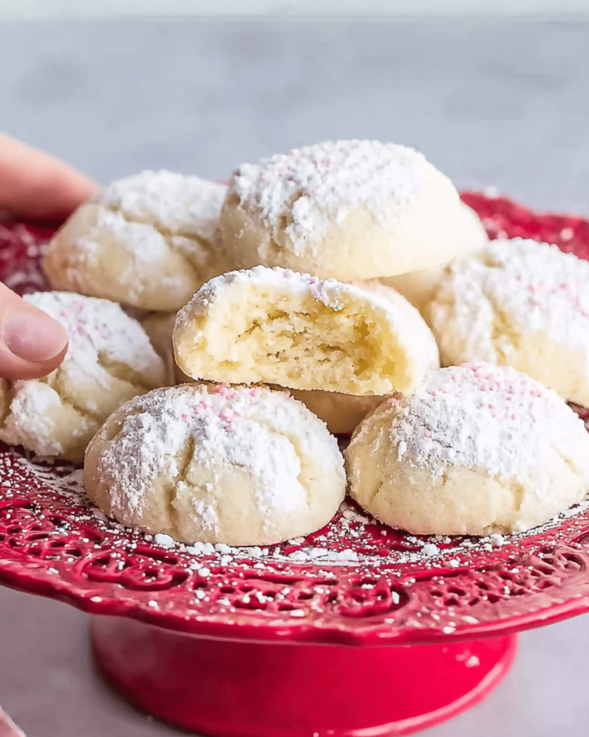 Melting moment cookies dusted with powdered sugar, displayed on a red decorative cake stand, with one cookie showing a soft, crumbly interior.