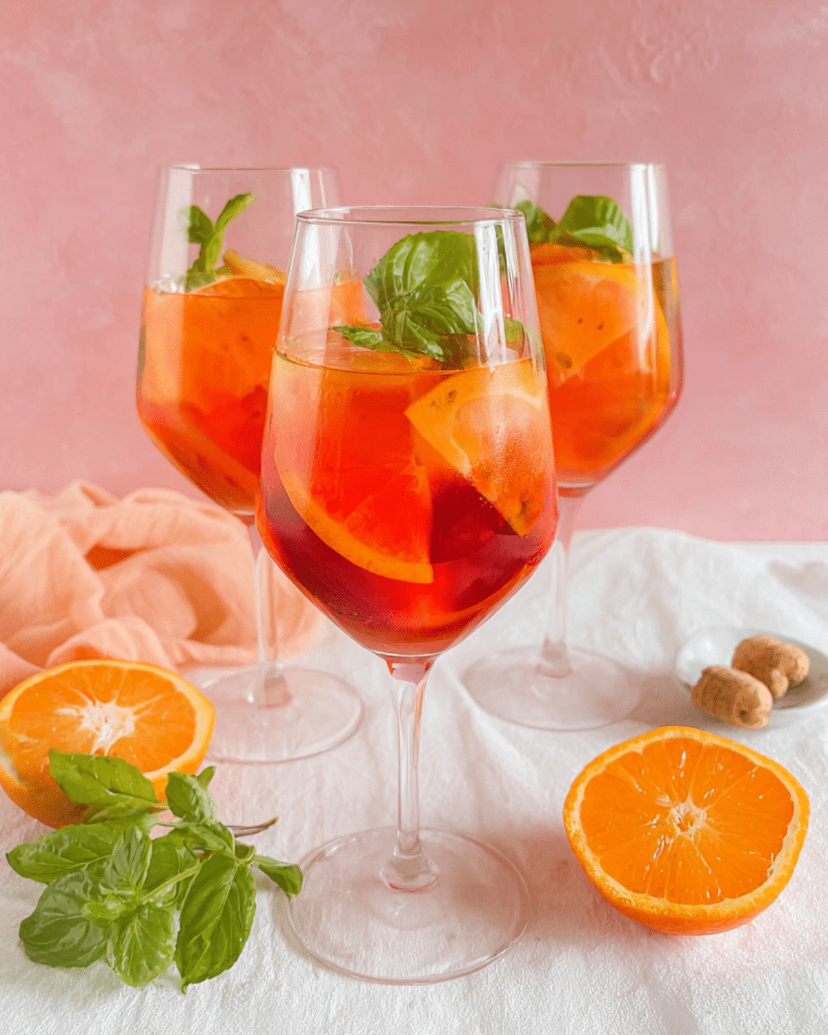 Rosé Aperol spritz cocktails served in wine glasses with orange slices and fresh basil, set against a pink background.