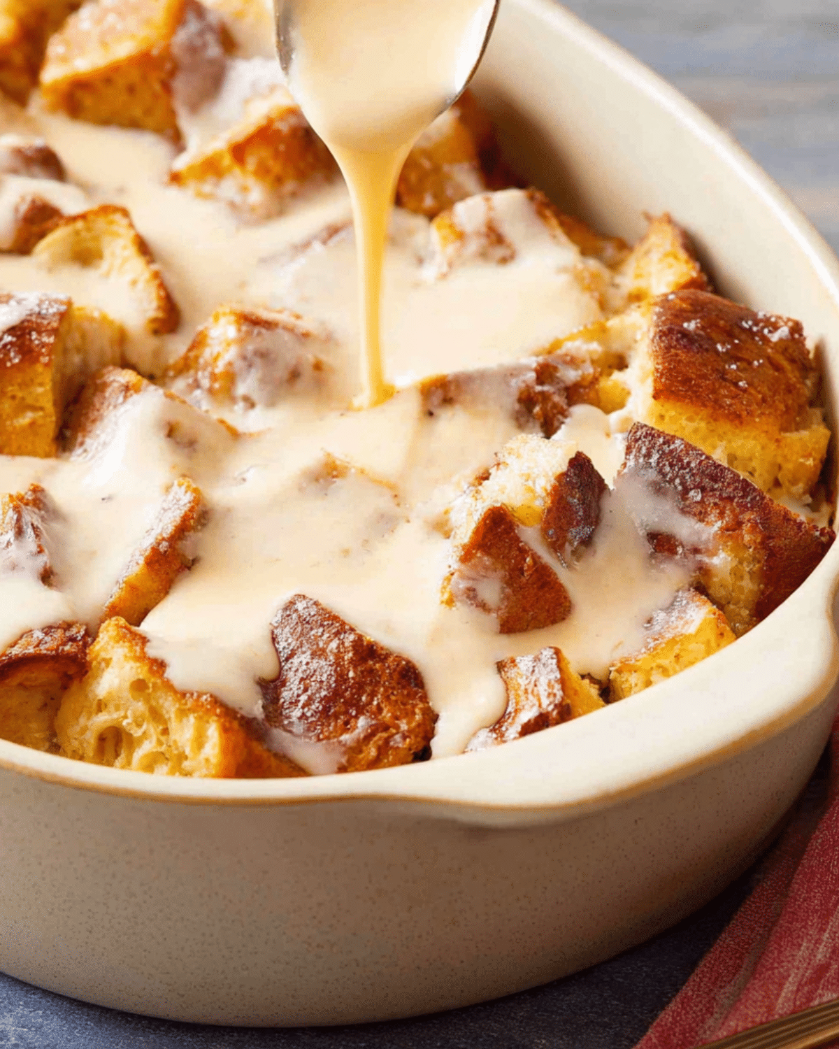 A baking dish filled with golden vanilla bread pudding, topped with warm vanilla sauce being drizzled from a spoon.