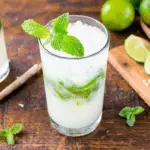 A glass of coconut rum mojito garnished with fresh mint, filled with crushed ice and muddled mint leaves, set on a wooden table with limes.
