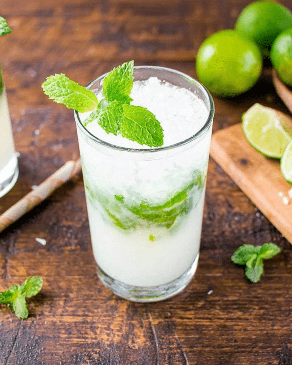 A glass of coconut rum mojito garnished with fresh mint, filled with crushed ice and muddled mint leaves, set on a wooden table with limes.
