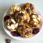 A bowl filled with homemade energy balls made from oats, chocolate chips, dried cranberries, and almond slices, with a few ingredients scattered around.