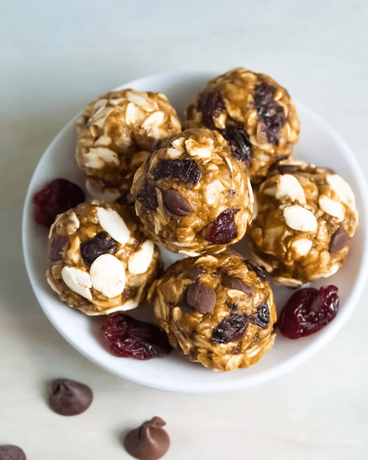 A bowl filled with homemade energy balls made from oats, chocolate chips, dried cranberries, and almond slices, with a few ingredients scattered around.