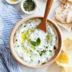 Healthy Tzatziki Sauce in a wooden bowl, topped with olive oil and fresh mint, surrounded by pita bread, herbs, and lemon slices.