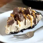 A slice of creamy No-Bake Snickers Bar Pie topped with chopped Snickers, chocolate drizzle, and caramel sauce on a white plate.