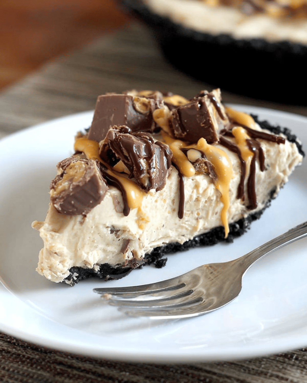 A slice of creamy No-Bake Snickers Bar Pie topped with chopped Snickers, chocolate drizzle, and caramel sauce on a white plate.