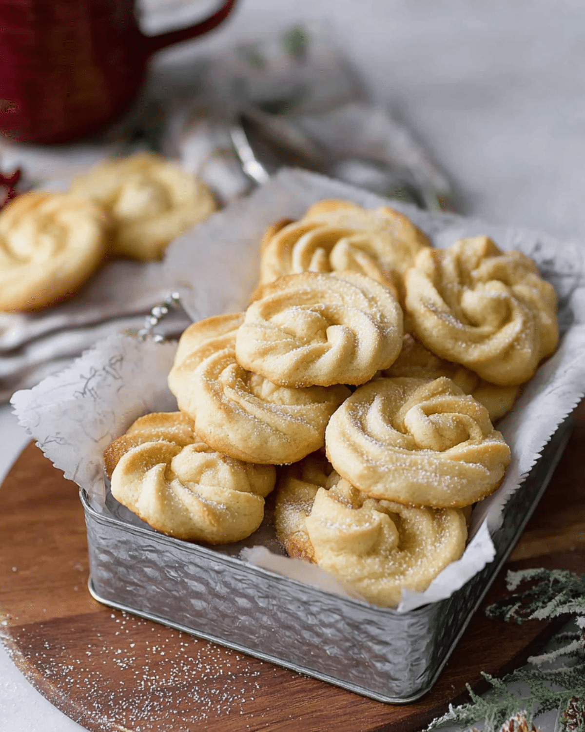 A tin filled with golden Danish butter cookies in a swirl shape, lightly dusted with sugar and arranged on parchment paper.