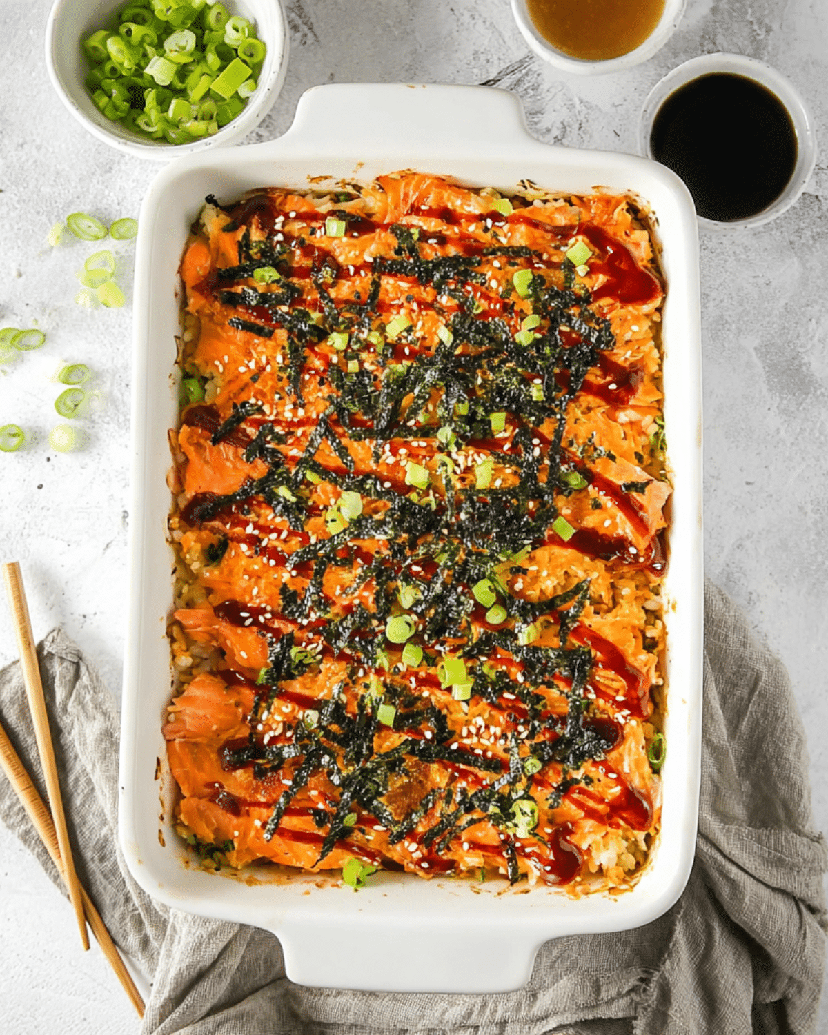 Spicy salmon sushi bake in a white baking dish, topped with seaweed strips, green onions, sesame seeds, and spicy sauce.